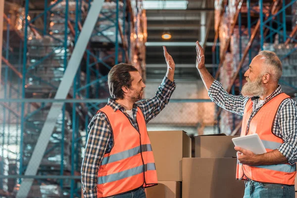 Side view of smiling warehouse workers giving high five while working with digital tablet